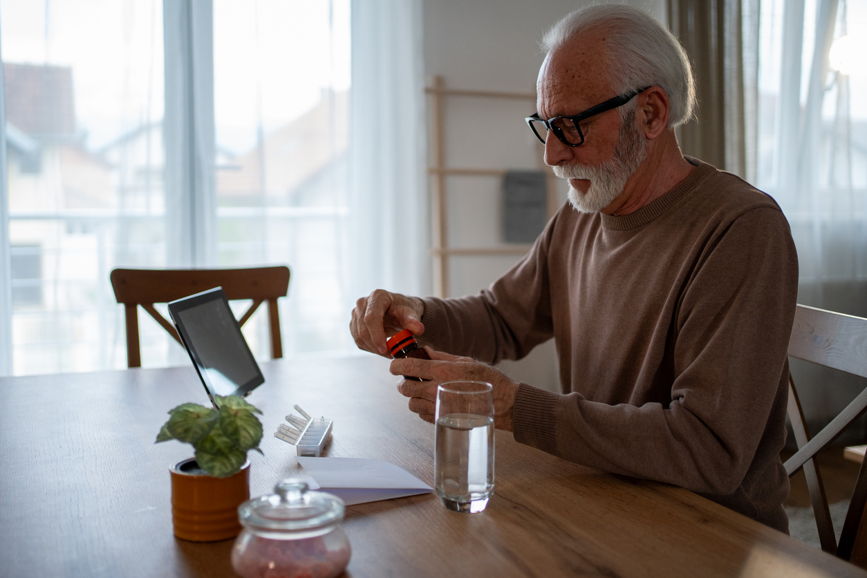 Elderly man opening a medicine bottle at home, preparing to take his pills while using a digital tablet and organized pill container on the table, emphasizing independent living and wellness