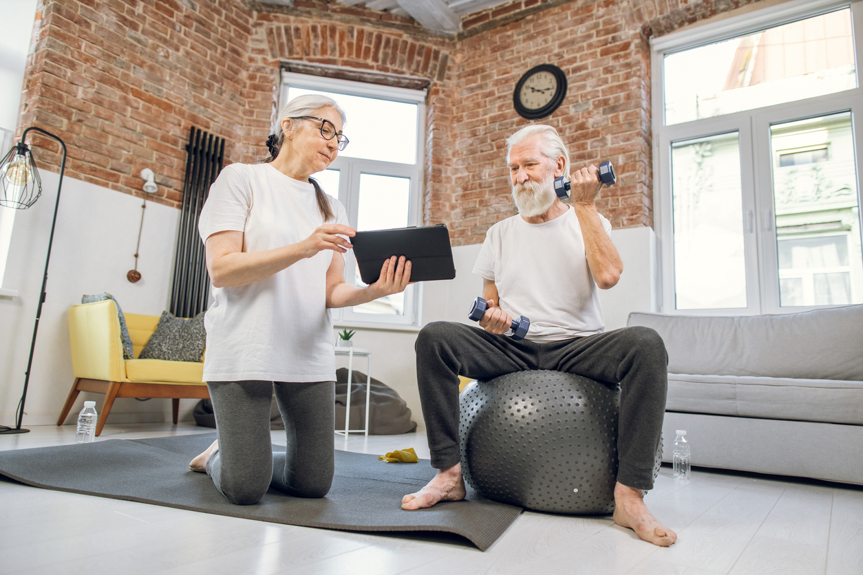 Aged woman holding digital tablet while standing on knees on yoga mat. Grey haired man sitting on fitball and training with dumbbells. Online tutorial for domestic workout.